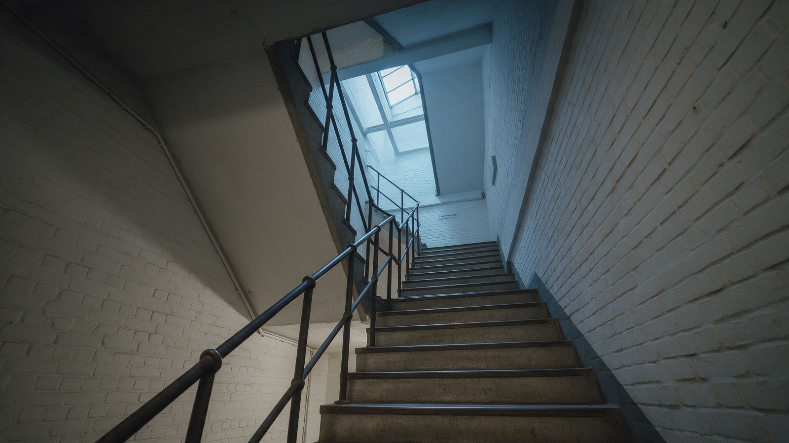 Betontreppe mit schwarzem Stahlgeländer, oben fällt Tageslicht durch ein Oberlicht